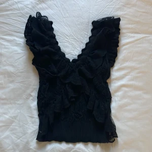 Svart spetsblus med volang från Zara - Supersöt svart blus från Zara med djup v-ringning och breda volangärmar. Toppen är i spets med virkade detaljer och har en croppad passform. Säljes i storlek S.