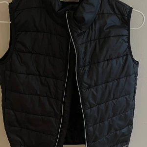 Svart dunväst från H&M - Svart quiltad dunväst från H&M med hög krage och dragkedja framtill. Västen har en stilren look med lätt glansigt material och diskreta sömmar i horisontella rader. Perfekt att slänga över en hoodie eller tröja för extra värme.