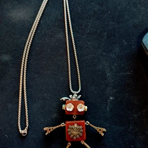 Robot-halsband med Eiffel-torn detaljer - Unikt halsband med en robotberlock i brunt och guld. Robotens kropp är dekorerad med blommor och ett hjärta med texten 'AMOUR'. Armar och ben har små Eiffel-torn och kugghjul som detaljer. Kedjan är i guldton och berlocken har en cool, lekfull design.