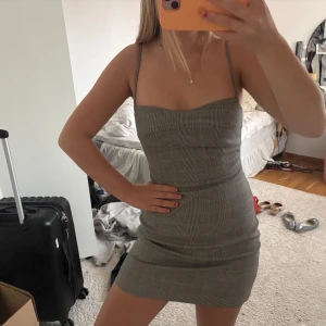 Grå rutig kortklänning med smala axelband - Säljer en grå rutig kortklänning med smala axelband och figurnära passform. Klänningen har en söt, hjärtformad urringning och är perfekt för dig som gillar en stilren och trendig look. Tunt och lätt material som känns skönt mot huden.