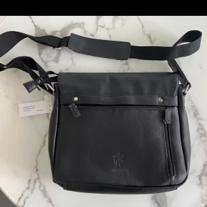 Svart axelväska från Leonhard Heyden - Snygg svart axelväska i skinn från Leonhard Heyden. Väskan har justerbar axelrem, flera dragkedjefack framtill och präglad logga. Perfekt storlek för vardagsprylar och stilren look. Passar dig som vill ha en clean och praktisk väska.