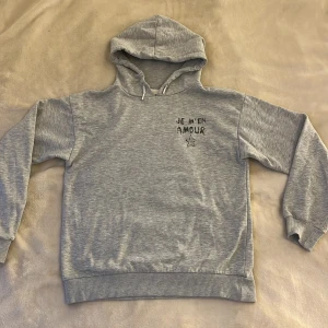Grå hoodie från Gina Tricot  - Grå hoodie från Gina Tricot med svart texttryck 'Je m'en amour' och en stjärna både fram och bak. Den är i storlek 146/152. Den är använd och är lite nopprig men det går att få bort.💓
