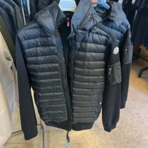 Svart Moncler pufferjacka med huva - Snygg svart pufferjacka från Moncler med quiltad framsida och ärmar i mjuk bomull. Jackan har huva, dragkedja och en cool ficka med dragkedja på ärmen. Perfekt för dig som vill ha en stilren och sportig look. Finns i alla storlekar 