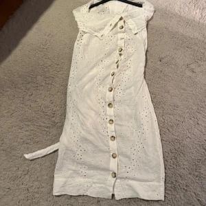 Vit broderad klänning AllSaints - Superfin vit klänning från AllSaints med broderade hålmönster, knappar framtill och knytband i midjan. Offshoulder-modell med krage och lätt A-linjeformad siluett. Perfekt för sommarens alla tillfällen!