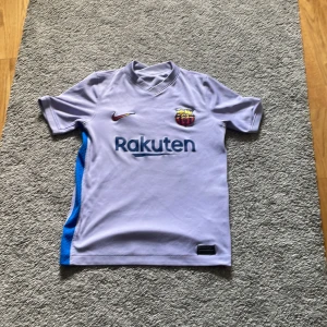 Ljuslila FC Barcelona Nike tröja - Säljer en ljuslila FC Barcelona fotbollströja från Nike med klubbmärke och Rakuten-tryck på bröstet. Tröjan har korta ärmar, blå och röda detaljer på sidorna samt Unicef-tryck på ryggen. Tillverkad i lätt och ventilerande material, perfekt för träning eller match.