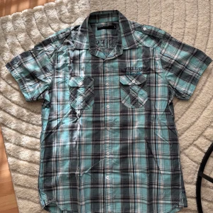 Rutig kortärmad skjorta från Identity - Snygg rutig skjorta från Identity i blått, svart och vitt. Skjortan har korta ärmar, två bröstfickor med lock och knappar samt broderad text på bröstet och ryggen. Perfekt för dig som gillar en avslappnad och cool stil.