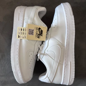 Nike Air Force 1 - Klassiska Nike Air Force 1 sneakers i helvitt skinn med perforerad tå, rund tå och platt sula. Snygg och tidlös modell med snörning och diskret Nike-logga på sidan. Perfekta för dig som vill ha en clean och fräsch look till vardags.