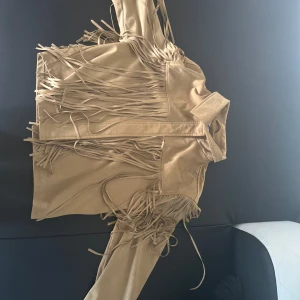 Beige mockajacka med fransar - Säljer en beige mockajacka med snygga fransdetaljer längs ärmar och rygg. Jackan har klassisk krage och stängs framtill.