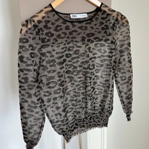 Leopardmönstrad meshblus från Zara - Transparent meshblus från Zara med leopardmönster i svart och grått. Blusen har rund halsringning, långa ärmar med resår vid ärmslut och midja för en puffig effekt. Material: 58% nylon 42% polyester 