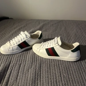 Gucci Ace vita sneakers med gröna detaljer - Gucci Ace sneakers i vitt skinn med klassiska gröna och röda ränder på sidorna och grön detalj i mockaimitation på hälen. Platt sula, rund tå och vita snören. Kommer med originalpåse, extra skosnören och äkthetsbevis. Perfekta för dig som vill sticka ut med stil. Pris kan diskuteras.