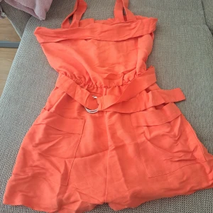 Orange jumpsuit shorts med bälte och fickor - Snygga orange jumpsuit shorts med hög midja och breda ben, tillverkade i ett lätt tyg. De har stora framfickor och ett matchande tygbälte med silverfärgad ringdetalj. Perfekta för varma dagar och ger en cool, avslappnad vibe.