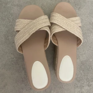 Beiga platåsandaler med flätade band - Snygga sandaler med breda, korslagda flätade band i beige textil och hög platåsula i repimitation. Mjukt beige innersula med vit detalj vid hälen. Perfekta för en chill sommarlook och ger extra höjd utan att tumma på komforten.