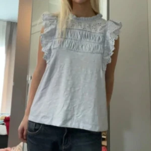 Ljusblå blus med volang och spets - Supersöt ljusblå blus med volangdetaljer på axlarna och spets upptill. Blusen är ärmlös med broderade detaljer och rund hals. 