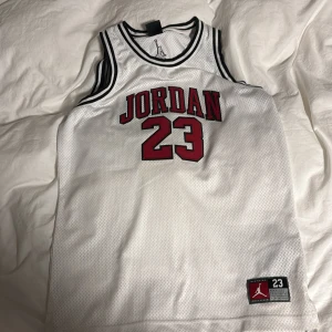 Vit Jordan basketlinne  - Säljer en vit basketlinne från Jordan med klassiskt rött och svart tryck, nummer 23 på bröstet och svarta detaljer vid hals och ärmhål. Perfekt för dig som gillar sportig stil och vill sticka ut med en ikonisk look.