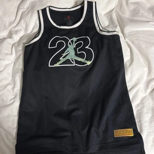 Jordan basketlinne svart  - Snyggt svart basketlinne från Jordan med stort 23-tryck och Jumpman-logga på bröstet. Vit kant runt ärmhål och halsringning samt guldfärgad Jordan-detalj nertill. Perfekt för sport eller streetwear.