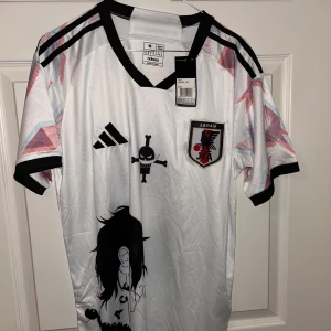 Japan Adidas fotbollströja One Piece M - Japan landslagströja från Adidas i vitt med svarta detaljer och coola One Piece-motiv fram och bak. Kortärmad modell med svart krage och ärmslut, samt grafiska mönster i rött och blått på axlarna. Perfekt för dig som älskar anime och fotboll.