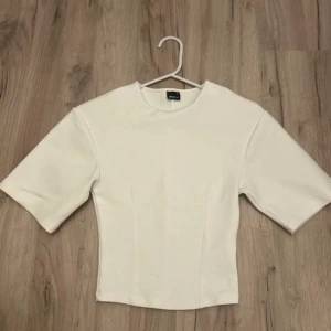 Vit croppad topp  - Säljer en stilren vit topp från Gina med croppad passform och vida, halvlånga ärmar. Toppen har en enkel design med rund halsringning och är gjord i ett mjukt bomullsmaterial. Perfekt för dig som gillar minimalistisk stil.