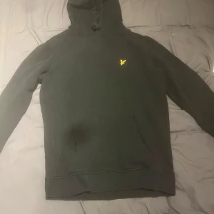 Svart hoodie från Lyle & Scott - Svart hoodie från Lyle & Scott med klassisk gul logga på bröstet. Tröjan har huva med snörning, stor magficka och långa ärmar. Stl XS
