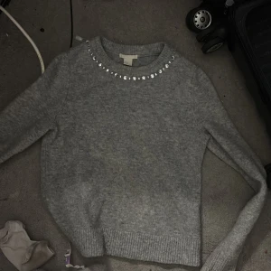 Grå stickad tröja med pärlor H&M XS - Supermjuk grå stickad tröja från H&M i storlek XS. Rund halsringning dekorerad med glittrande pärlor för en extra touch. Långärmad modell med ribbade muddar och nederkant. Perfekt för dig som gillar stilrena men ändå unika detaljer.