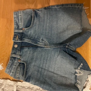 Blå jeansshorts med fransar - Snygga blå jeansshorts med fransiga kanter. Perfekta för sommardagar. De har en dragkedja och knapp framtill för enkel stängning.