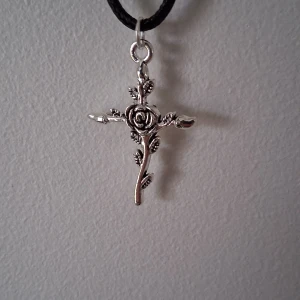 Silverfärgat halsband med roskors - Unikt silverfärgat halsband med ett korsformat hänge dekorerat med en vacker ros och blad. Hänget hänger på en svart snodd och ger en elegant touch till din stil.
