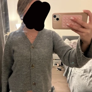 Grå kofta med knappar - Mysig grå kofta med knappar framtill. Perfekt för kyligare dagar och enkel att matcha med olika outfits. Koftan har en klassisk design med långa ärmar.