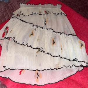 Blommig kjol från Ganni - Säljer en söt blommig kjol från Ganni. Kjolen är vit med färgglada blommor och har volangdetaljer med svarta kanter. Perfekt för en somrig look! Det står ingen storlek på men den är resår och har band så man kan välja det som passar än själv. Men jag har storlek S och den passar mig så skulle säga att den är u storlek S/M