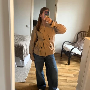 Beige trenchcoat från Vila - Säljer en snygg beige trenchcoat från Vila. Den är aldrig använd men har ingen lapp kvar. Storlek 38. Jackan har en dubbelknäppt design med stora svarta knappar och klassiska slag. Den har långa ärmar med justerbara remmar vid ärmsluten och en rak passform. Perfekt för vår och höst!