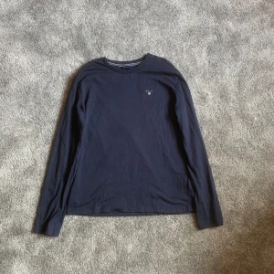 Mörkblå långärmad tröja från GANT - Säljer en stilren mörkblå långärmad tröja från GANT med broderad logga på bröstet. Perfekt för en avslappnad och snygg look. Tröjan är tillverkad i mjukt material och har en klassisk passform.