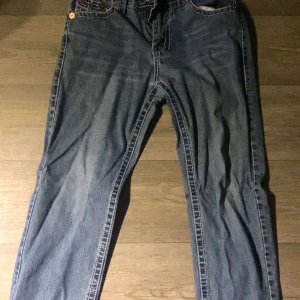 True Religion Jeans - Snygga blå jeans med unika broderade detaljer på bakfickorna. De har en klassisk femficksdesign och en knappgylf. Perfekta för en avslappnad stil.