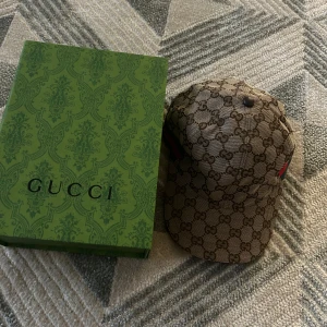 Beige keps från Gucci - Snygg beige keps från Gucci med det klassiska GG-mönstret. Kepsen har en grön och röd randdetalj på sidan och är tillverkad i Italien. Perfekt för att ge din outfit en lyxig touch.