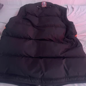 Väst från Parajumpers - Säljer en svart storlek L dunväst från Parajumpers med dragkedja och praktiska fickor. Västen har en stilren design och är perfekt för kyligare dagar. Den har även orangea detaljer som ger en snygg kontrast. Skriv vid funderingar