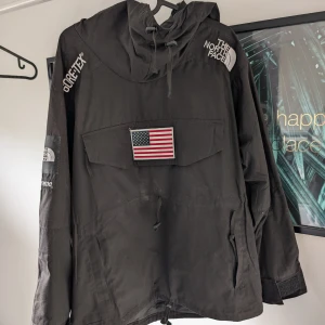 Svart jacka från The North Face x Supreme - Säljer en svart jacka från The North Face i samarbete med Supreme. Jackan har en stor amerikansk flagga på bröstet och ryggen, samt flera loggor inklusive GORE-TEX. Den har en justerbar huva och dragkedja framtill.