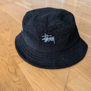 Svart bucket hat från Stussy - Snygg svart bucket hat från Stussy med broderad logga framtill. Perfekt för en avslappnad stil. Tillverkad i mjukt material för bekväm passform.
