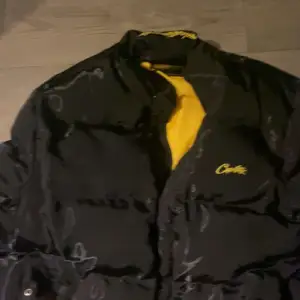 Säljer en svart pufferjacka från Corteiz med gul insida och broderad text på kragen. Jackan har en cool design med dragkedja och är perfekt för kyligare dagar. Den har en modern look och är både stilren och funktionell.