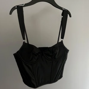 Svart korsett från her[body] - Snygg svart korsett från her[body] med justerbara axelband och formande design. Perfekt för att ge en stilren och elegant look. Tillverkad i ett glansigt material som ger en lyxig känsla.