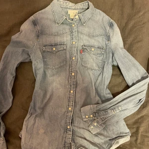 Blå jeansskjorta från Levi's - Säljer en klassisk blå jeansskjorta från Levi's med knappar framtill och två bröstfickor. Skjortan har långa ärmar och en stilren design som passar perfekt till vardags. Tillverkad i Sri Lanka.