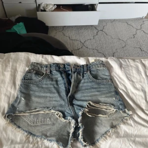 Blå jeansshorts med slitningar - Säljer ett par blå jeansshorts med coola slitningar och fransar. De har en klassisk femficksdesign och är perfekta för en avslappnad stil. Passar bra till sommarens alla äventyr!