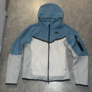 Blå och vit jacka från Nike - Säljer en snygg blå och vit jacka från Nike med dragkedja och huva. Jackan har en cool design med en ficka på ärmen och svarta detaljer. Perfekt för en sportig look!