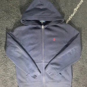 Mörkblå hoodie från Ralph Lauren - Säljer en snygg mörkblå hoodie från Ralph Lauren med dragkedja och en liten röd logga på bröstet. Perfekt för en avslappnad stil. Den har långa ärmar och en praktisk huva.