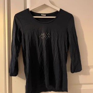 Mörkblå topp från Atelier GS - Snygg mörkblå topp från Atelier GS med trekvartsärmar och ett glittrigt mönster framtill. Perfekt för att lägga till lite bling i vardagen. Tillverkad i ett bekvämt material som passar bra till olika tillfällen.