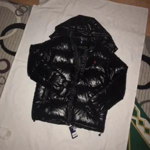 Svart dunjacka från Ralph Lauren - Säljer en snygg svart dunjacka från Ralph Lauren med huva och dragkedja. Jackan har en glansig finish och är perfekt för kyligare dagar. Den har en klassisk design med en liten logotyp på bröstet. Perfekt för dig som vill hålla stilen i vinter! 🖤