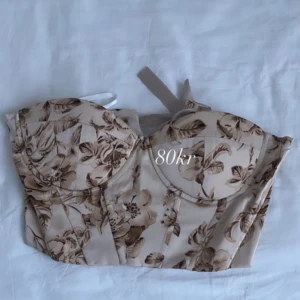 Beige blommig korsettopp - Säljer en snygg beige korsettopp med blommigt mönster. Toppen har en strukturerad design med vadderade kupor och är axelbandslös. Perfekt för att ge en elegant touch till din outfit.