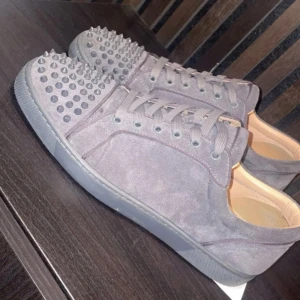 Grå sneakers med nitar - Snygga grå sneakers med nitar på tån och röd sula. Skorna har snörning och är tillverkade i mocka. Perfekta för att ge din stil en edgy touch.