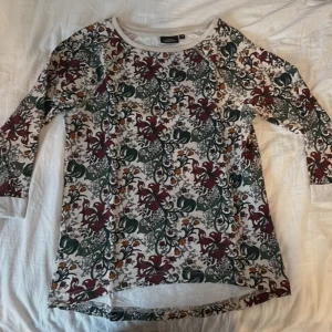 Mönstrad långärmad tröja från KappAhl - Säljer en mönstrad långärmad tröja från KappAhl i storlek XS. Tröjan har ett blommigt mönster i grönt, vinrött och orange på en vit bakgrund. Perfekt för en färgglad och stilren look.