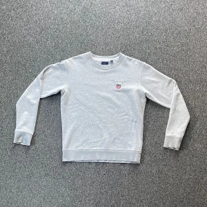 Grå sweatshirt från GANT - Säljer en stilren grå sweatshirt från GANT med broderad logotyp och emblem på bröstet. Tröjan har rund hals och långa ärmar, perfekt för en avslappnad stil. Passar bra till både jeans och chinos.