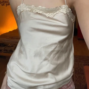 Vit satinlinne med spetsdetaljer - Säljer ett stilrent vitt linne i satin med fina spetsdetaljer längs kanten. Perfekt för en elegant look. Axelbandslös design som ger en chic touch.