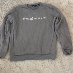 Grå långärmad från Sail Racing - Säljer en stilren grå sweatshirt från Sail Racing med tryck på bröstet. Tröjan har långa ärmar och en rund halsringning. Perfekt för en avslappnad stil.