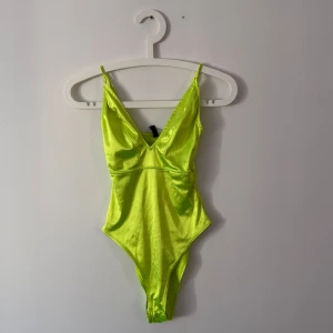 Neongrön bodysuit - Snygg neongrön bodysuit med smala axelband och djup v-ringning. Tillverkad i ett glansigt material som ger en cool look. Perfekt för att sticka ut!