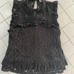Svart spetsblus med volangdetaljer - Säljer en elegant svart spetsblus från Voyelles med vackra volangdetaljer. Blusen är ärmlös och har en fin knäppning i nacken. Perfekt för att ge en stilfull touch till din outfit. Det står också på den att det är storlek M men skulle säga att den passar mer som XS/S. Den är absolut väl använd men absolut inte i något dåligt skick, kom gärna privat för frågor eller fler bilder 😊